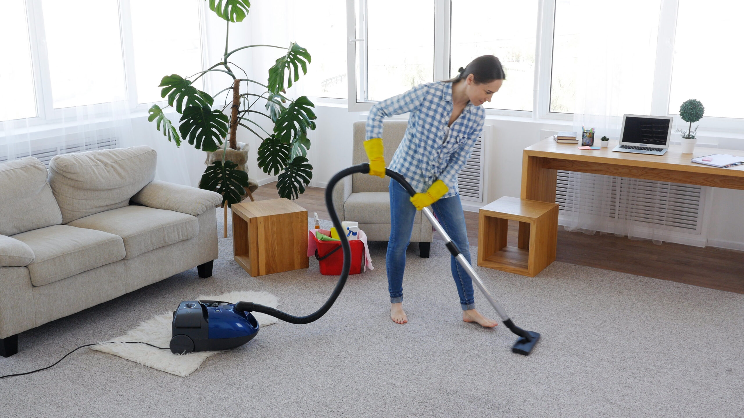 A woman wearing yellow cleaning gloves and a blue plaid shirt is vacuuming a light-colored carpet in a bright living room. A blue and black vacuum cleaner is visible on the floor. This is an example related to Albuquerque Area Rug Cleaning by Bear Oriental Rug Cleaning.