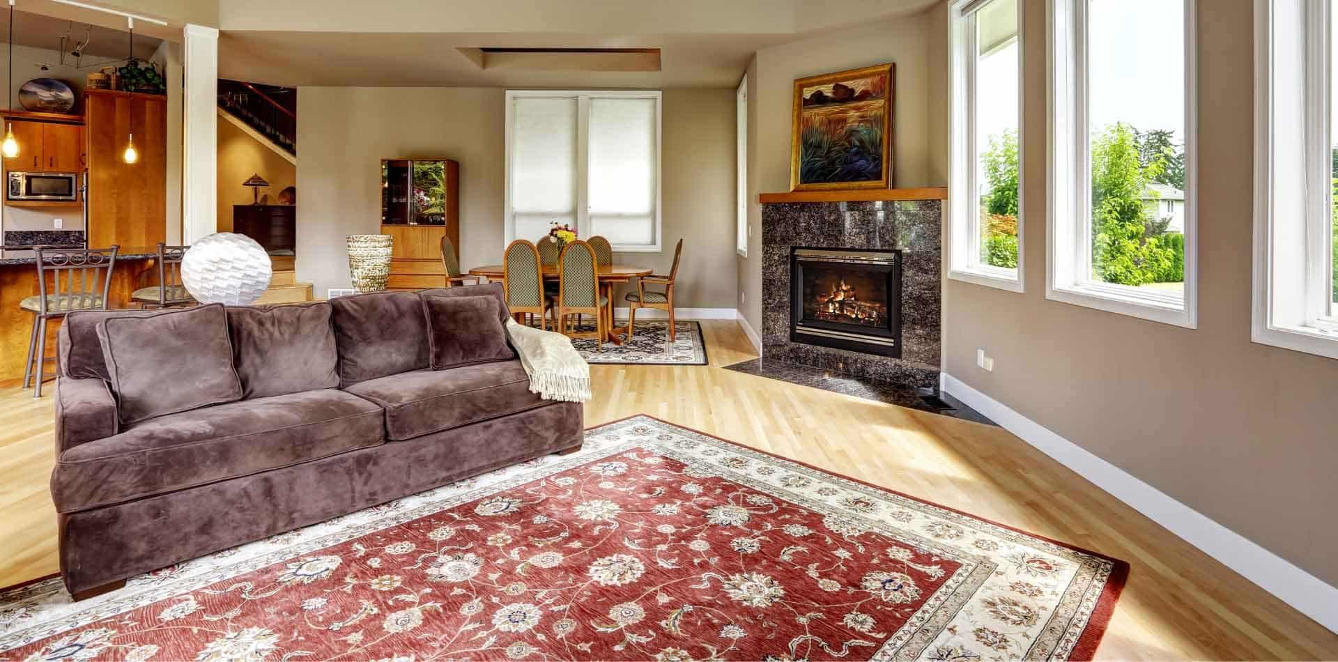 A luxurious living space with light wood floors, a dark brown sofa, and a large, patterned red oriental rug. A fireplace with a dark marble surround is visible, along with a dining area in the background. This is an example of a setting that requires Albuquerque Area Rug Cleaning by Bear Oriental Rug Cleaning.
