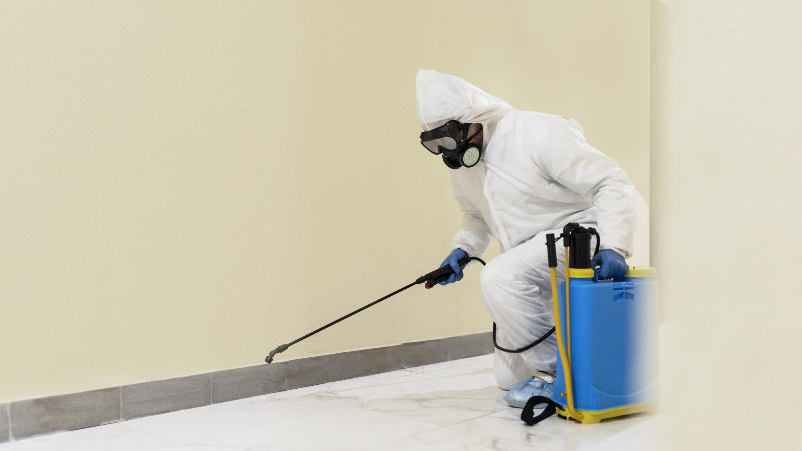 Albuquerque Mold Mitigation specialist from Free Mold Inspections New Mexico kneeling on a tiled floor, wearing a white protective suit and respirator, applying a solution to a baseboard area using a backpack sprayer during mold treatment.