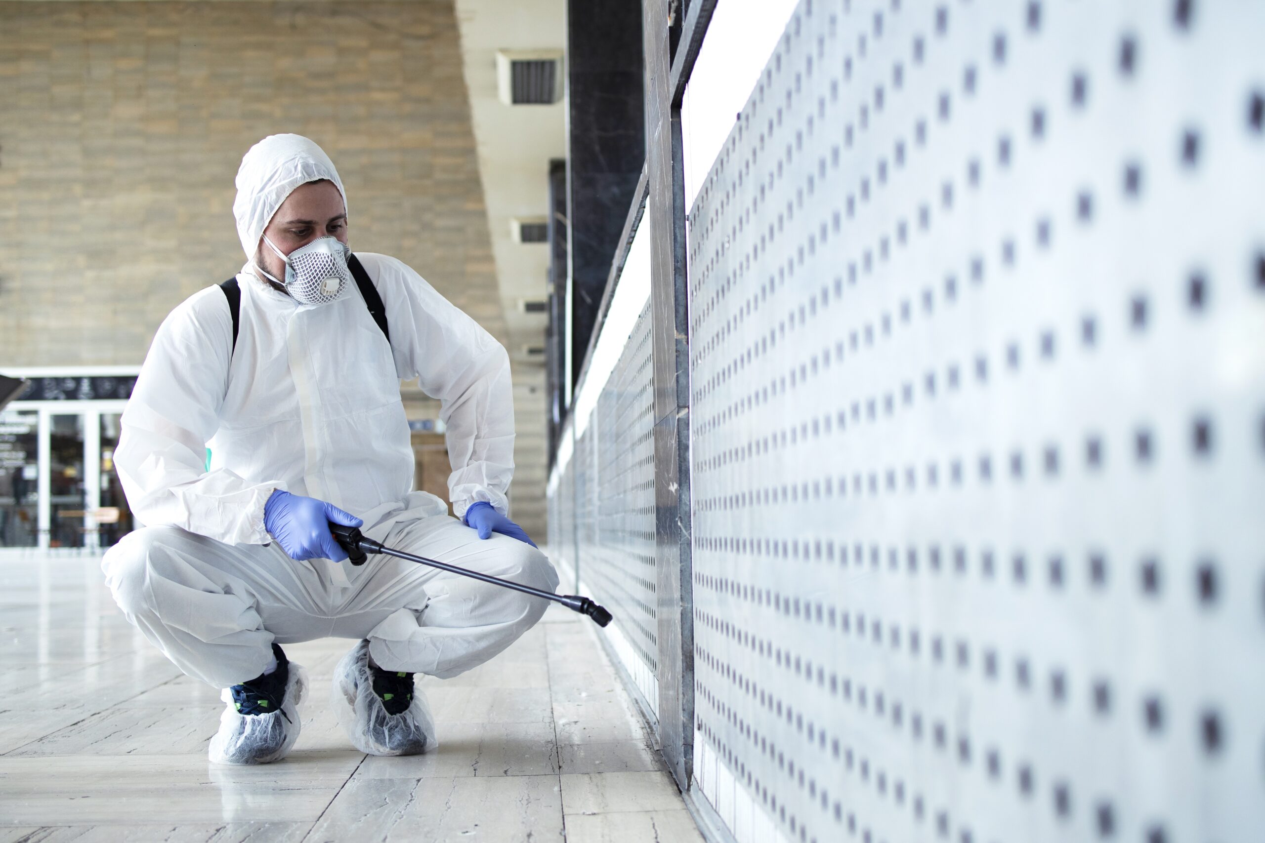 A professional technician in a white protective suit, respirator mask, gloves, and shoe covers is kneeling while spraying a solution on a surface with small holes.