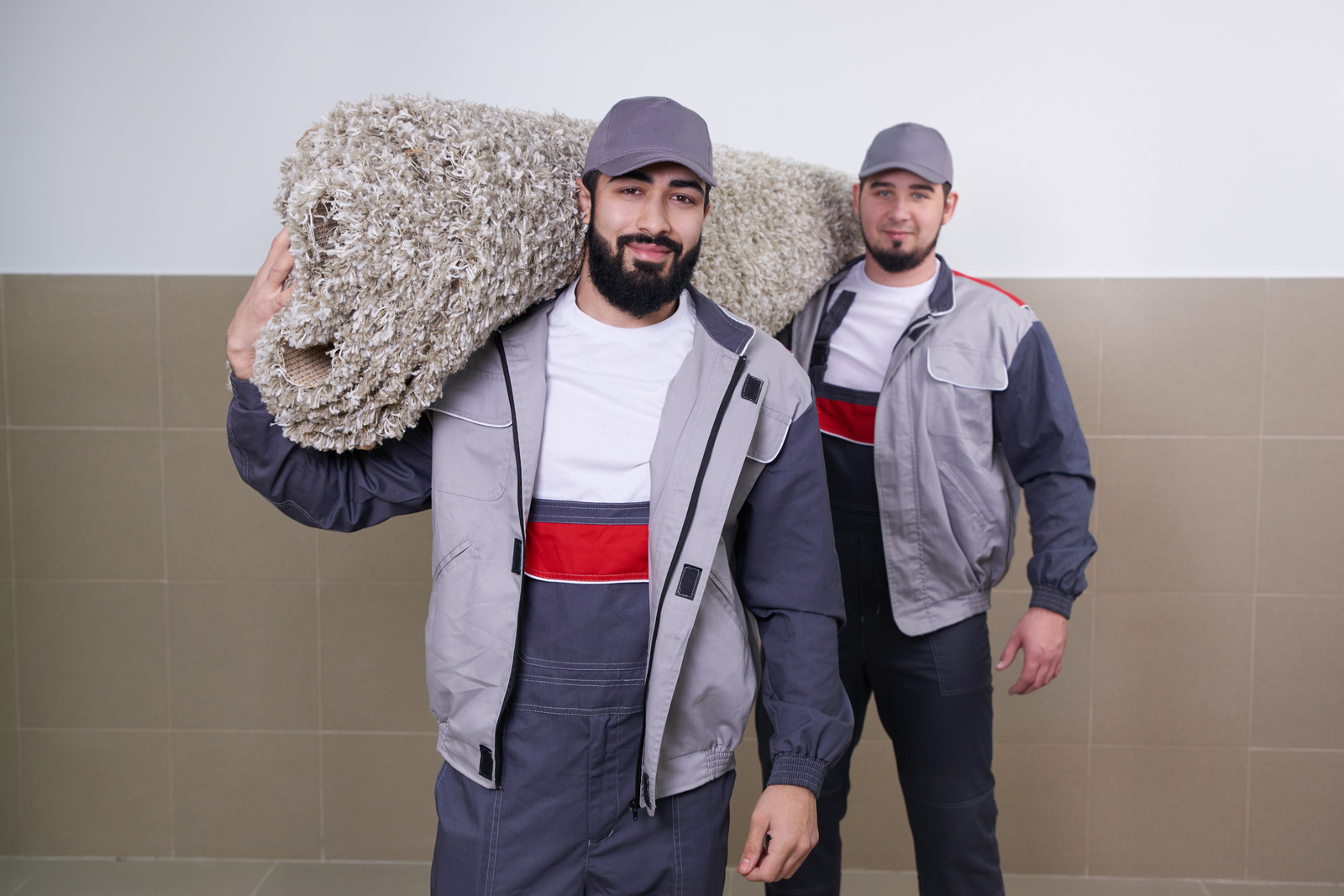 Two uniformed employees of Albuquerque Rug Cleaning Companies by Bear Oriental Rug Cleaning, one carrying a large rolled-up shag rug over his shoulder, stand ready to provide rug cleaning services.