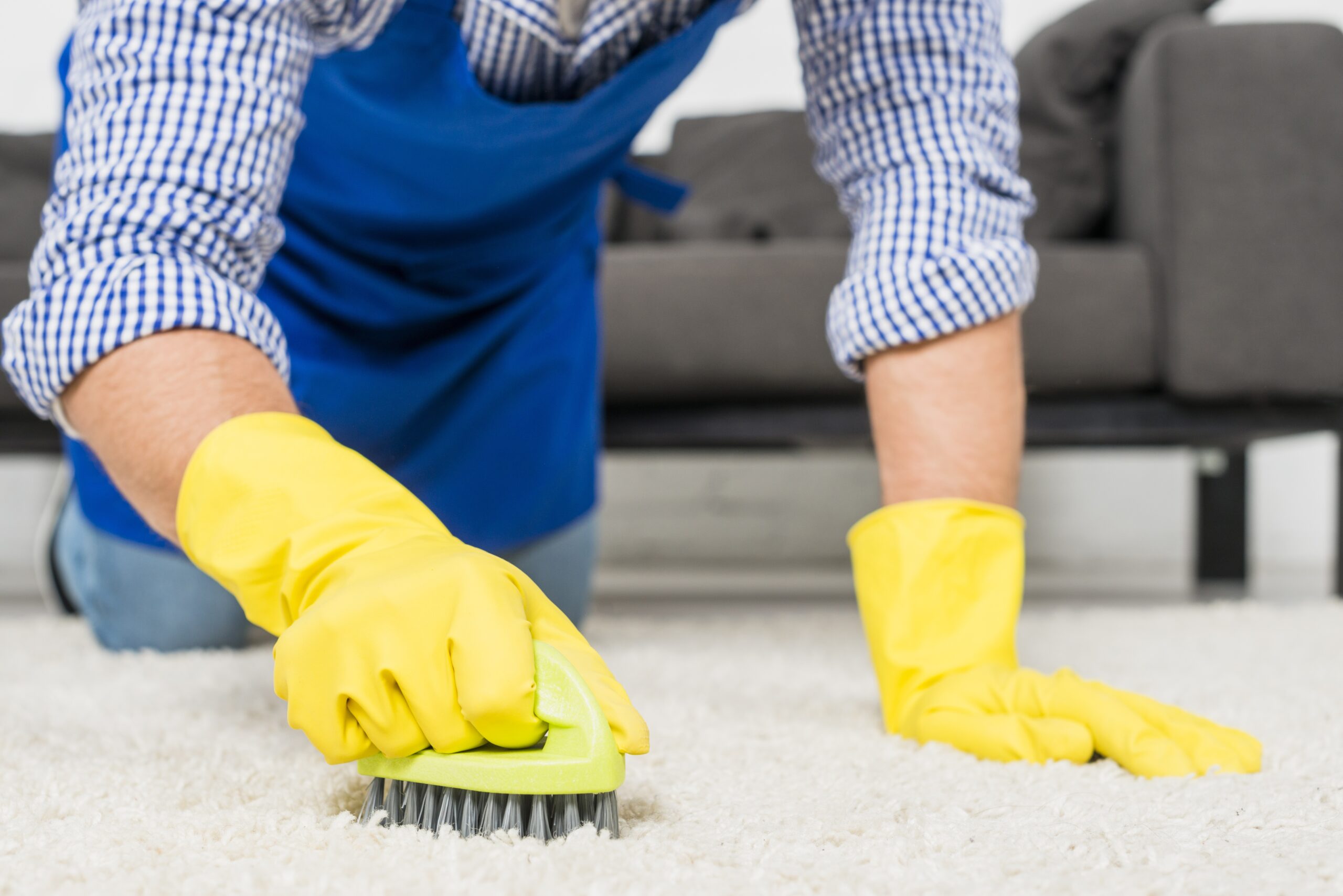 A close-up shot of a man's hands in bright yellow gloves kneeling on a white shaggy rug. He is scrubbing the rug with a green-handled brush. This image promotes the detailed work of Albuquerque Rug Cleaning Service by Bear Oriental Rug Cleaning.