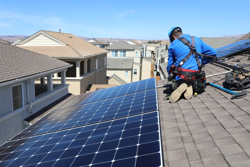 An installer wearing safety gear kneels on a residential shingle roof to work on newly installed solar panels in a suburban neighborhood, representing Asheville NC Residential Roofing Services by Burish Builder Asheville.