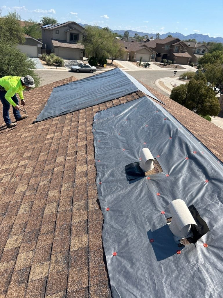 A worker from Burish Builder Asheville, an Asheville NC Roofing Company, prepares a brown shingled roof for replacement, installing a gray synthetic underlayment near two roof vents in a sunny suburban neighborhood.