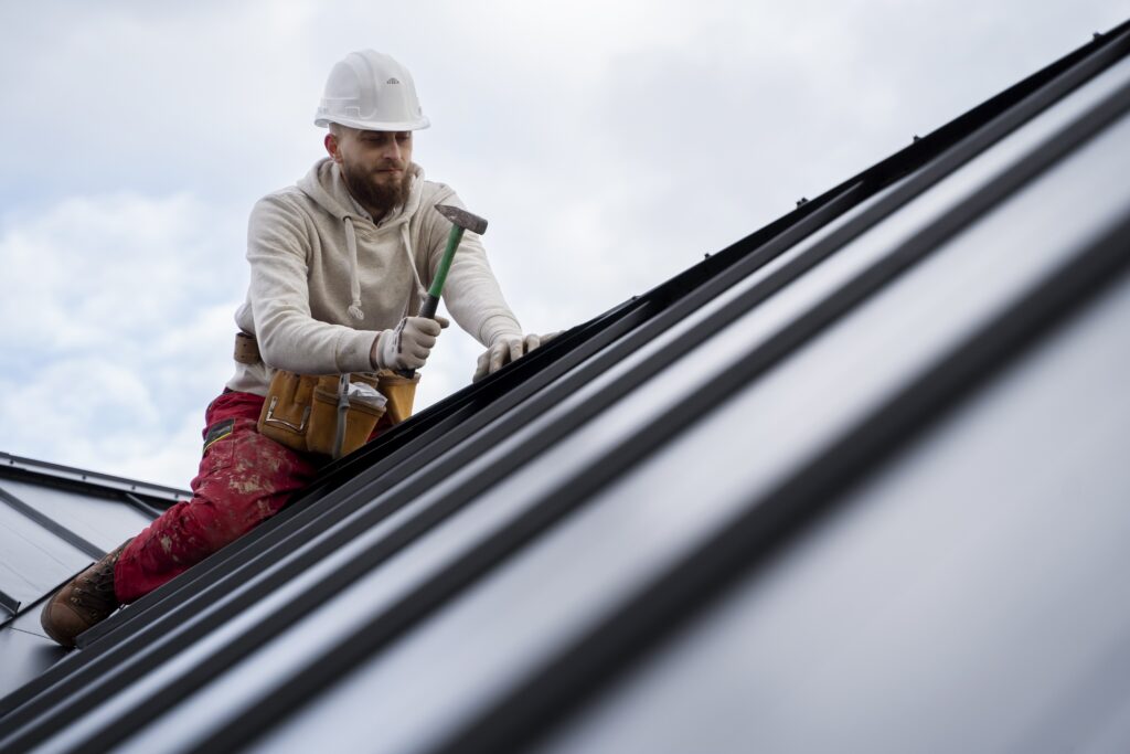 A close-up, low-angle shot of a bearded construction worker in a white hard hat and hoodie, with a tool belt, carefully installing Billings Metal Roofing by Wegner Roofing & Solar on a steep roof pitch, holding a hammer.
