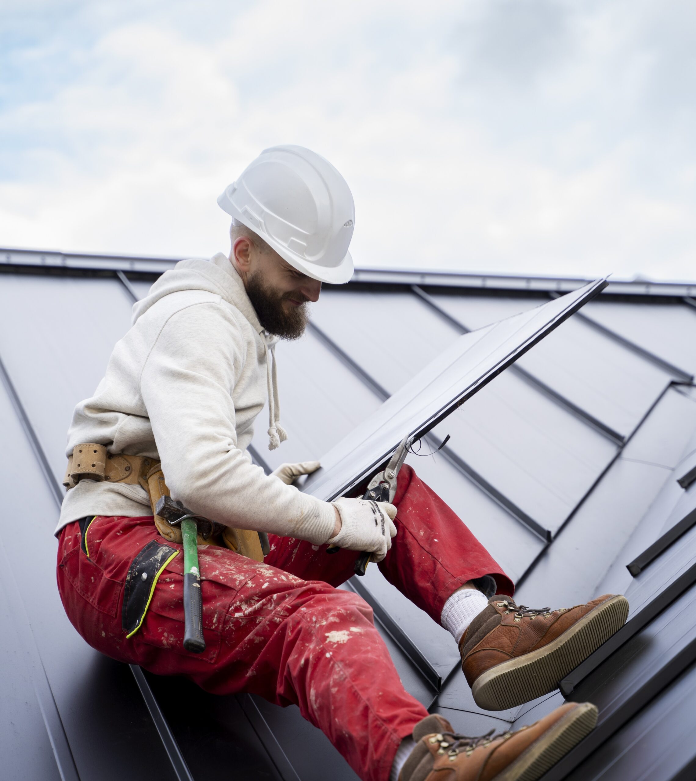 A contractor in a white hard hat and red pants, sitting on a black metal roof, using metal snips to cut a section of the Billings Metal Roofing panel for installation, representing Wegner Roofing & Solar.