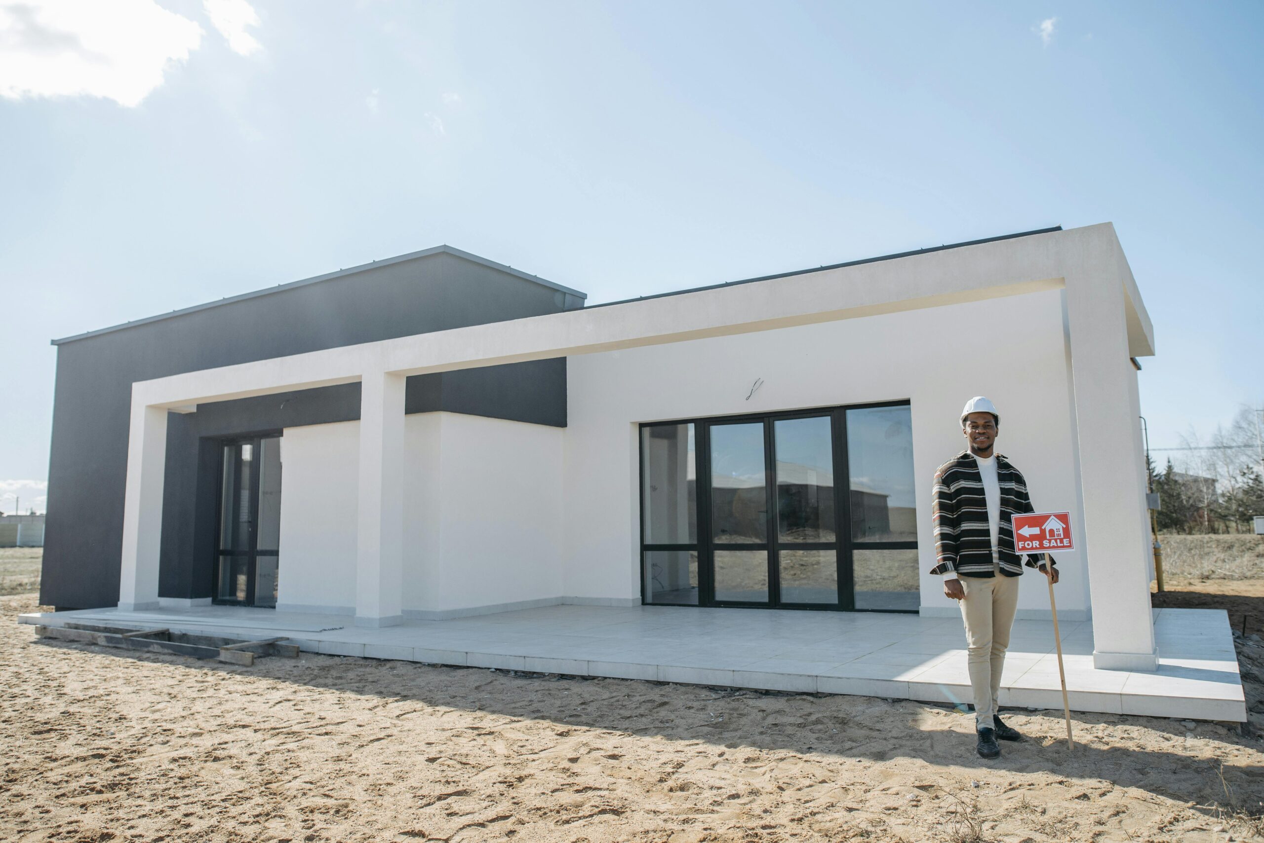 A smiling African-American male wearing a construction hard hat and a patterned shirt stands in front of a modern, white and black single-story house holding a red "FOR SALE" sign. This is a Billings Realtor photo by Living in Billings.