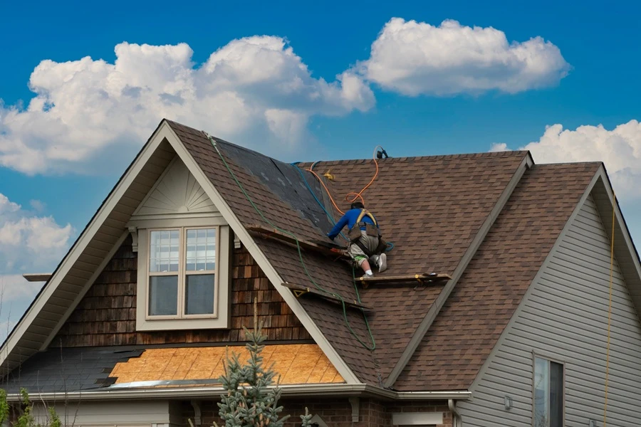 Bismarck Emergency Roofing Services by Wegner Roofing & Solar performing roof repair or installation on a residential home with brown shingles, showing one worker on the roof under a blue, cloudy sky.