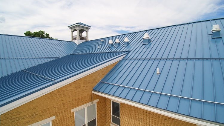 A vibrant blue standing seam Bismarck Metal Roofing system installed by Wegner Roofing & Solar on a building with yellow brick and white trim. The metal roofing has vertical ribs and various roof vents visible.