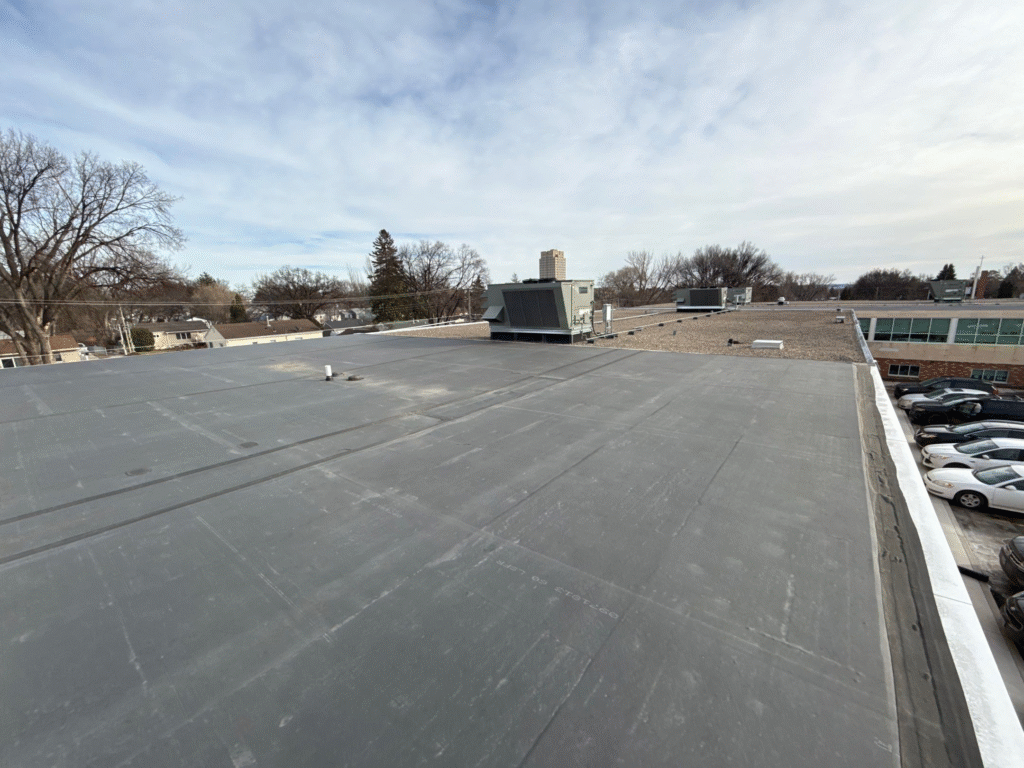 Wide view of a large, flat commercial roof, likely a Bozeman Commercial Roof installed by Wegner Roofing & Solar, covered with dark gray membrane material. A large HVAC unit is near the center, and a parking lot is visible on the right.