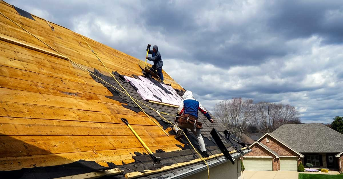 Two roofers working on a residential roof replacement, tearing off old shingles and installing insulation and underlayment, showcasing Charlotte NC Residential Roofing Services by Burish Builder CLT under a dramatic, cloudy sky.