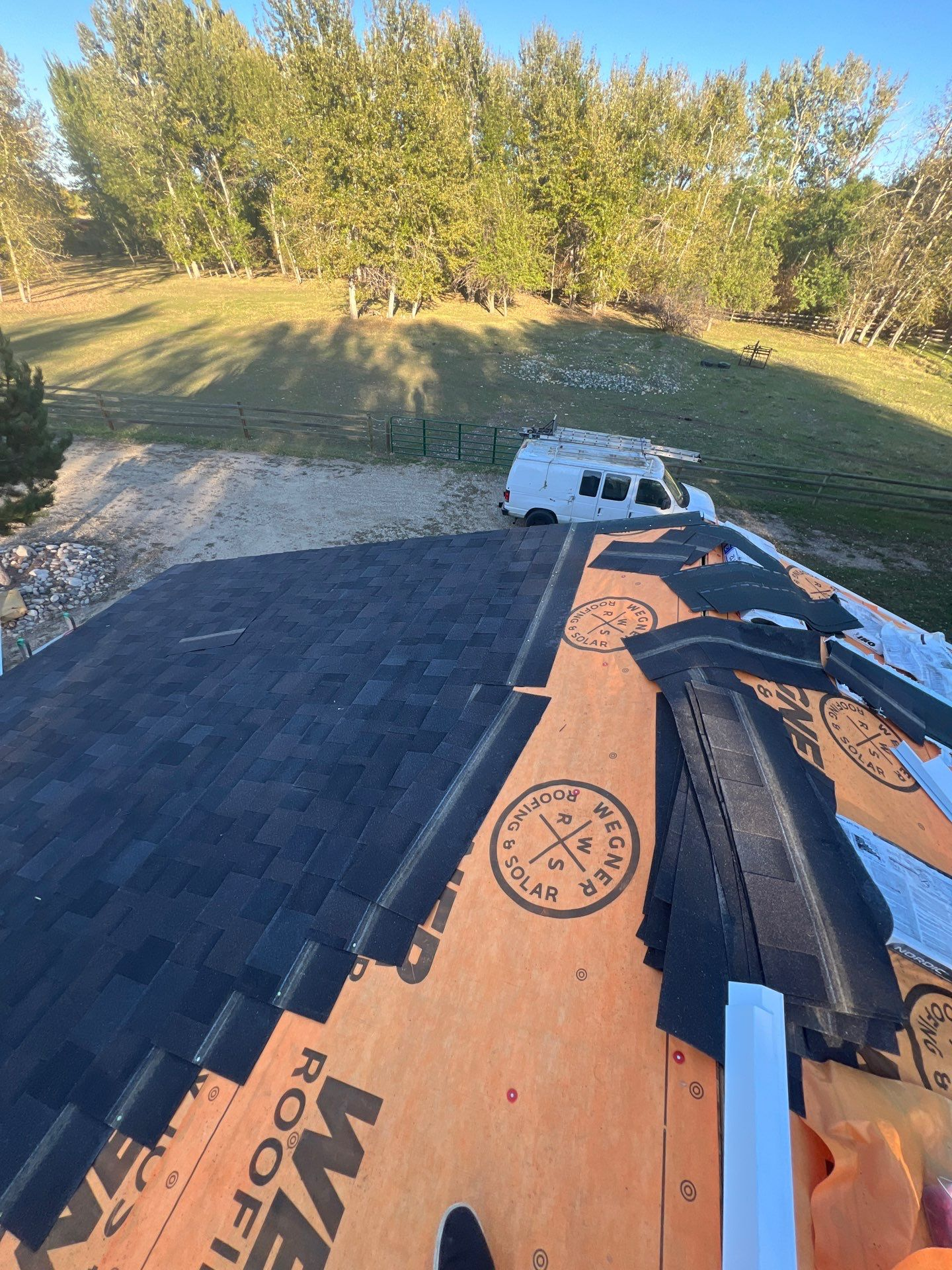 Dark asphalt shingle roofing installation in progress, showing new shingles laid over orange underlayment with the 'Wegner Roofing & Solar' logo visible, and a white work van parked in the background near a wooded area.