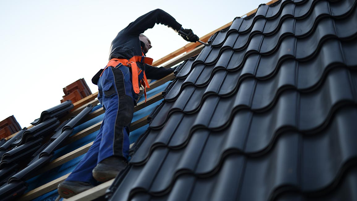 A roofer wearing an orange safety harness works on installing black metal roof tiles on a steep roof pitch. Edmond OK Commercial Roof Replacement by Roof One.