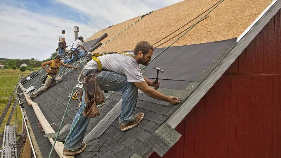 A crew of roofers from Roof One performing Edmond OK Residential Roof Repair. One roofer in the foreground is kneeling, hammering a nail into the new gray asphalt shingles, with exposed roofing felt and plywood sheathing visible above him. Other crew members are working on the steeper sections of the roof.