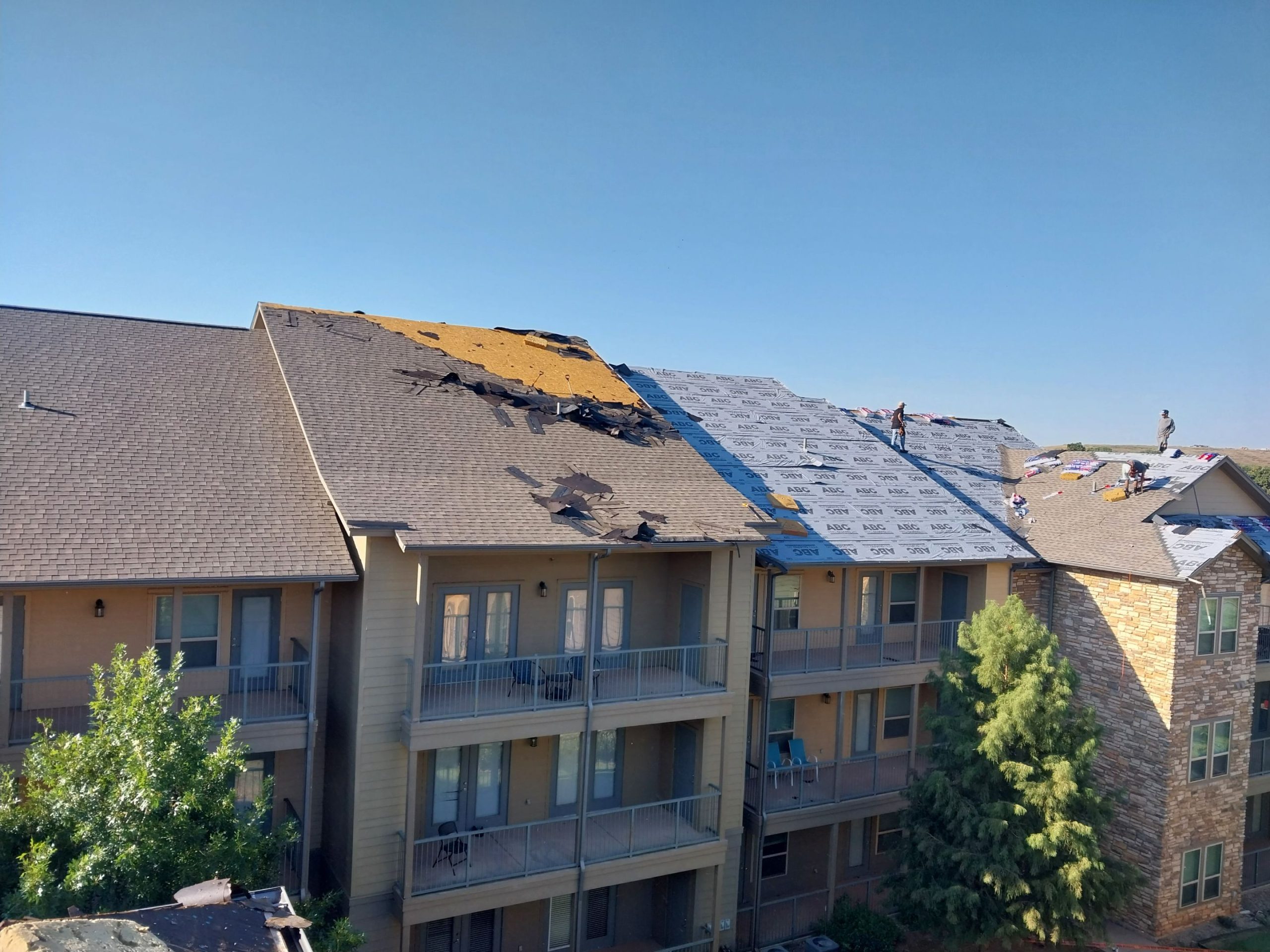 Wide shot of a large apartment complex roof being replaced as part of an Edmond OK Residential Roof Replacement by Roof One. The image shows sections of old roofing removed, exposing yellow decking, and new blue synthetic underlayment being installed by workers on a sunny day.