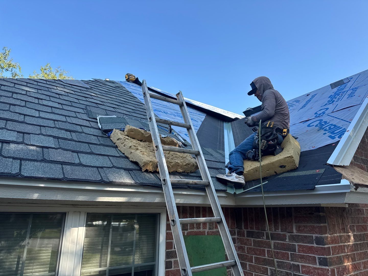 Edmond OK Roof Installation Services by Roof One: A close-up view of a roofer on a sunny day sitting on the hip of a roof, working on shingle installation next to a ladder and a roll of insulation, demonstrating the roofing process.