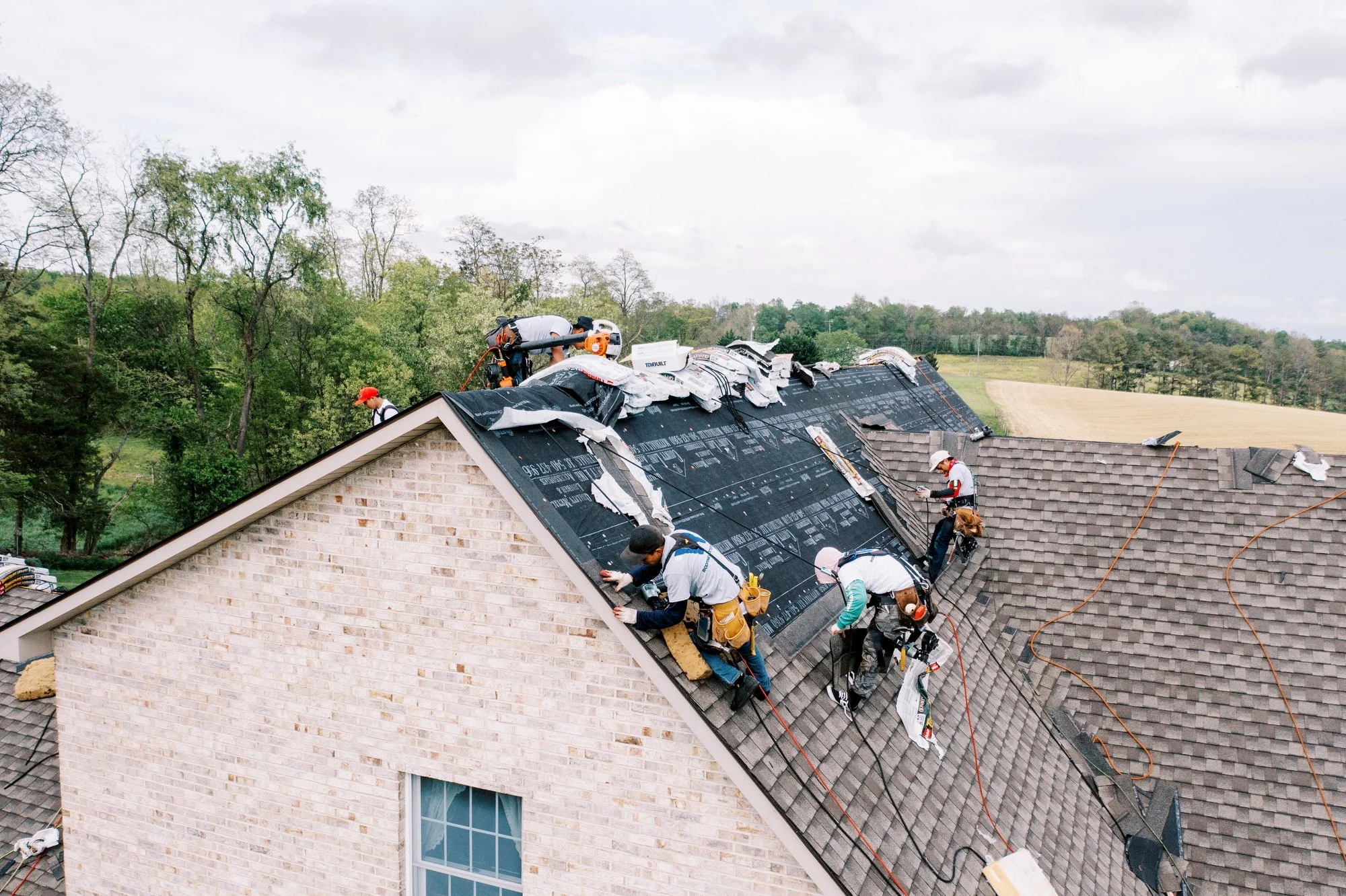 Edmond OK Roof Installation Services by Roof One: An elevated view of a team of roofers actively working on a large residential roof, installing new dark-colored shingles and underlayment, with a background of trees and countryside.