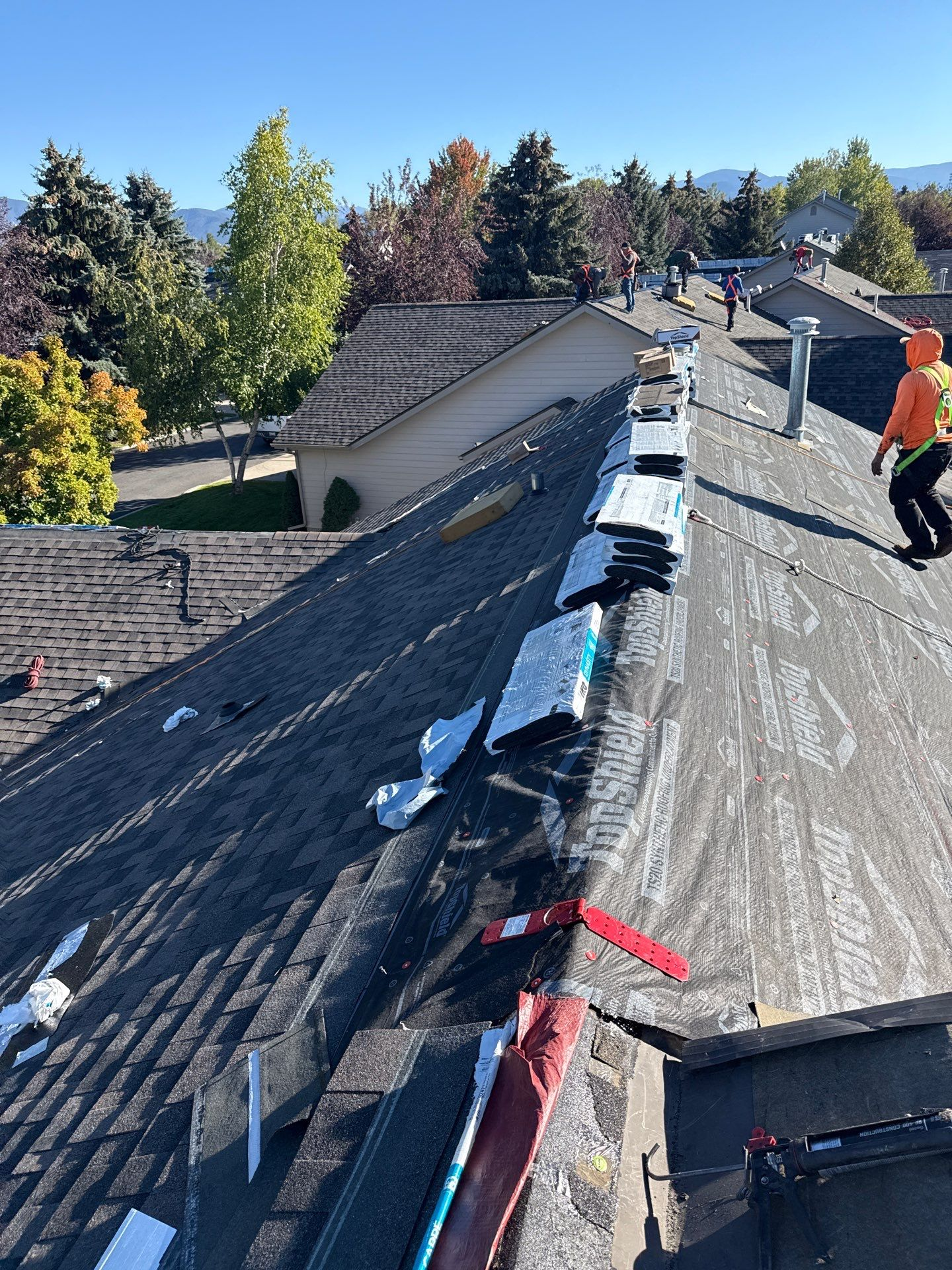 Roofing crew installing a new shingle roof, with materials staged along the ridge and workers in safety gear visible against a backdrop of trees and a mountain range. The work-in-progress shows exposed synthetic underlayment. Project by Forrest City NC Roofing Company by Burish Builder Asheville.