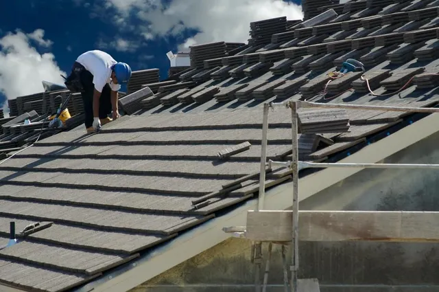 A roofer in a white shirt and blue hard hat works on installing gray shingles on a roof, with stacks of shingles nearby and scaffolding to the right, depicting a Forrest City NC Roofing Replacement by Burish Builder Asheville.