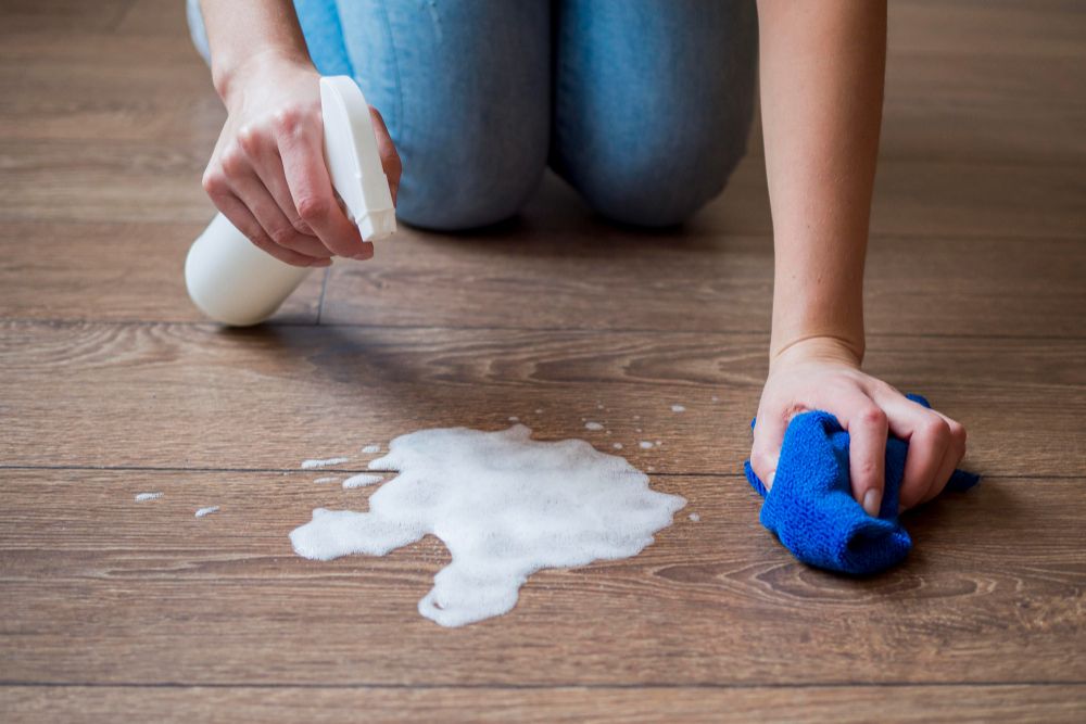 Removing wax buildup from hardwood floor with spray cleaner and blue cloth in Greensboro hardwood wax removal service.
