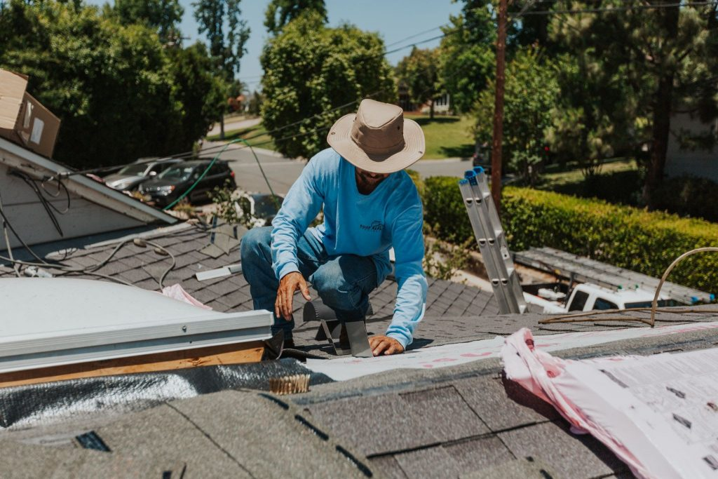 A roofer wearing a wide-brimmed hat and a blue long-sleeve shirt is crouched down working on a gray shingled roof, with tools and materials visible. This image captures a professional providing Hendersonville TN Residential Roofing Services by Burish Builder TN.