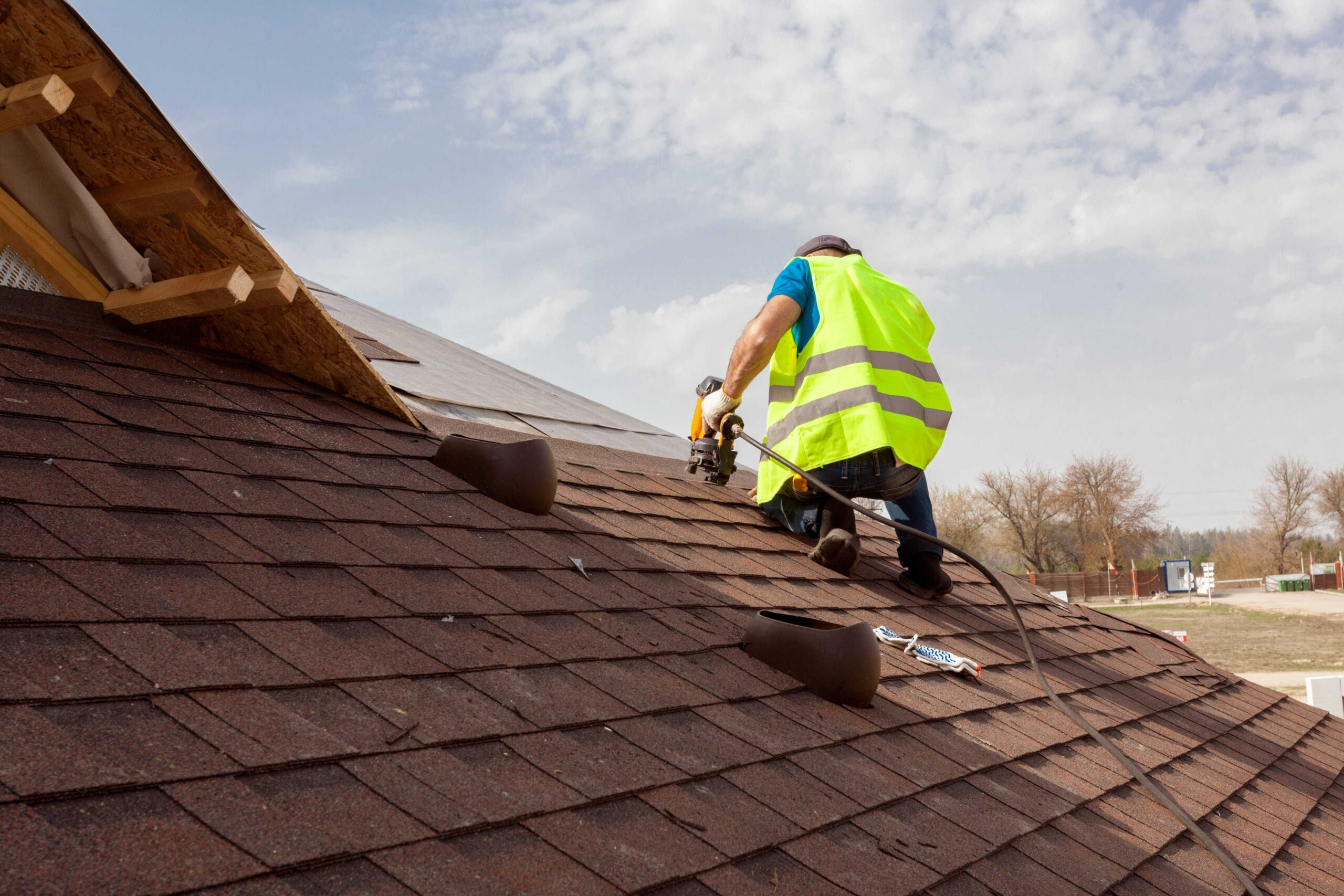 A roofer in a high-visibility vest using a nail gun to install brown asphalt shingles on a roof. Hendersonville TN Roofing Installation by Burish Builder TN.