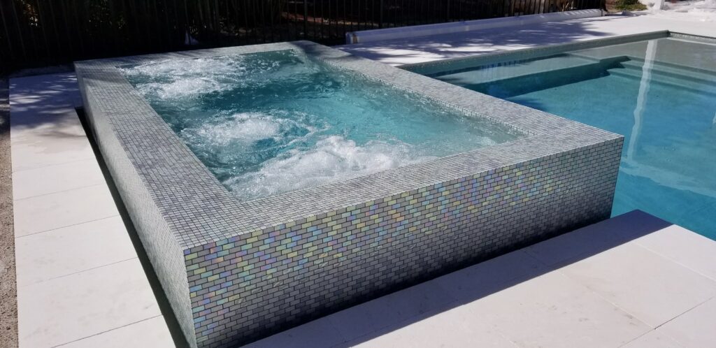 A rectangular, elevated spa with white-to-iridescent mosaic tiling on the exterior, showing active jets and bubbles, next to a larger swimming pool. This is a view of a Mooresville Above Ground Pool Installation by Craft Master Pools and Spas.