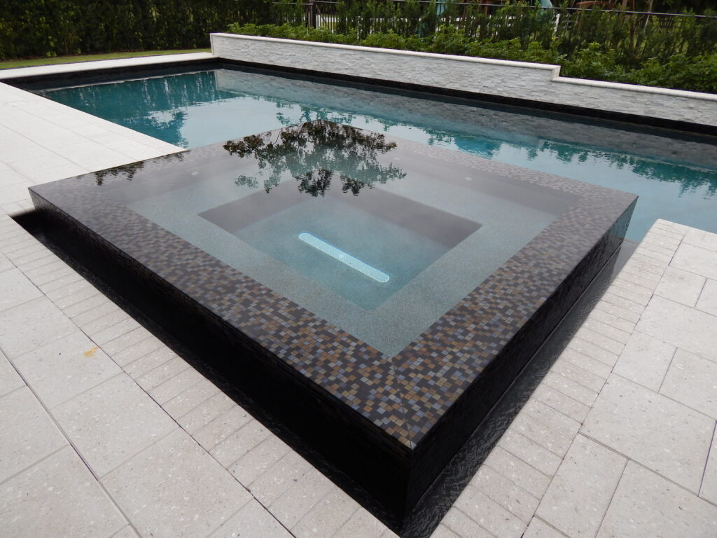 An elevated square spa with dark, iridescent mosaic tile trim, reflecting the surroundings, set against a larger pool with surrounding light-colored coping. This is a detail of a Mooresville Above Ground Pool Installation by Craft Master Pools and Spas.