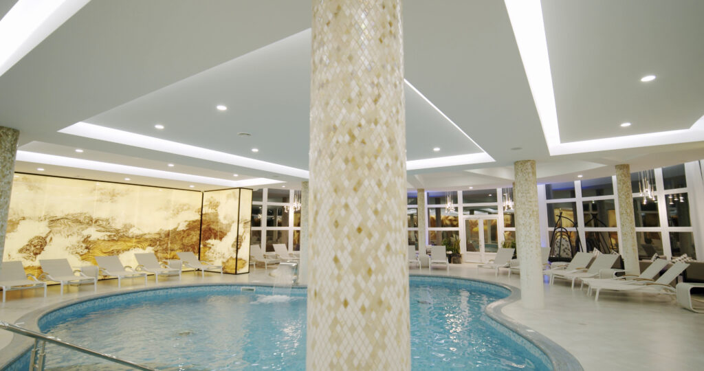 An elegant indoor, free-form swimming pool inside a bright, luxurious spa. The room features a large, central pillar covered in mosaic tiles, numerous white lounge chairs, and modern chandeliers, showcasing a high-end design by Craft Master Pools and Spas by Mooresville Pool Construction.
