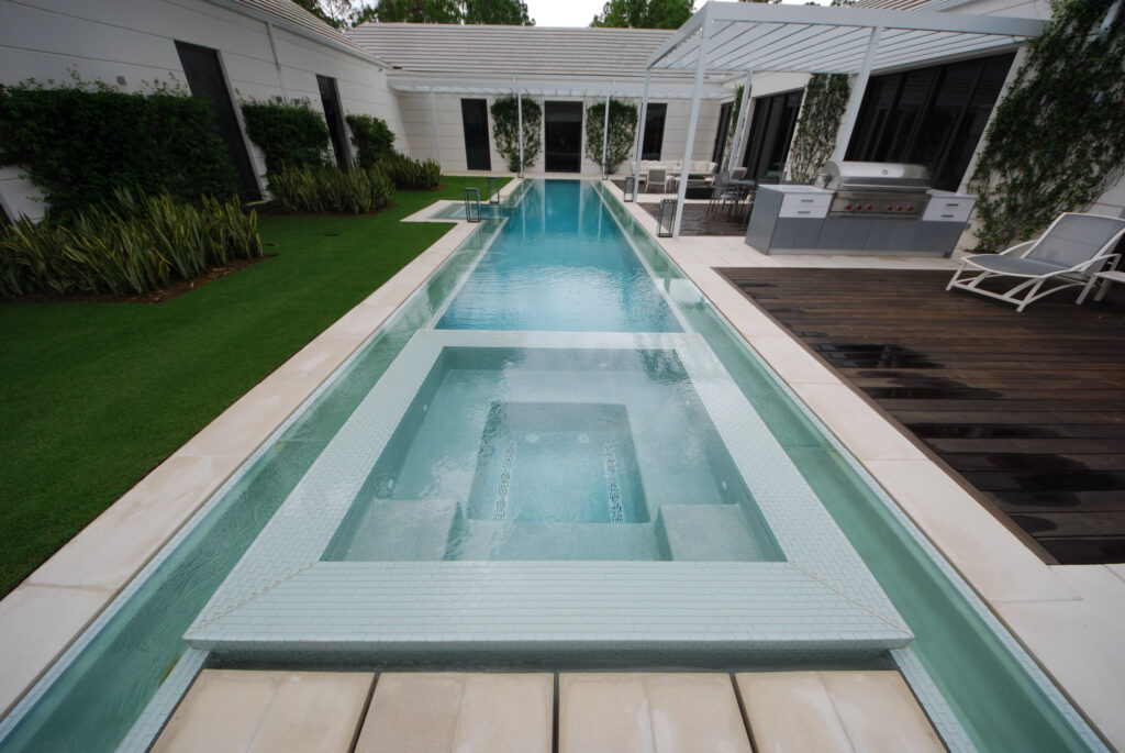 A sleek, modern rectangular lap pool with a raised, square-tiled spa in a luxurious backyard setting. This stunning project by Craft Master Pools and Spas by Mooresville Pool Construction features a unique glass wall design, a manicured lawn, and a dark wood deck with an outdoor kitchen.