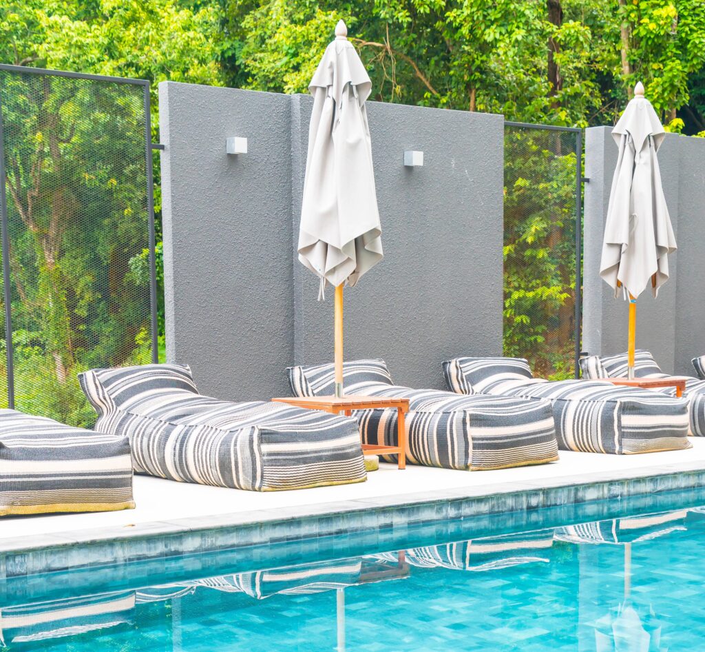 Luxurious poolside lounging area with modern grey wall, two closed beige umbrellas on wooden poles, and multiple striped bean-bag style lounge chairs next to a bright blue swimming pool, featuring a Mooresville Pool Renovation by Craft Master Pools and Spas.