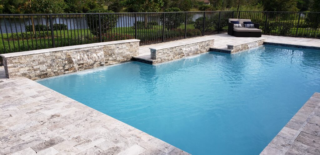 Stunning view of a newly renovated rectangular swimming pool with light blue water, surrounded by light-colored travertine coping, with two cascading waterfall features built into stacked stone walls, set against a fenced yard overlooking a pond, showcasing a Mooresville Pool Renovation by Craft Master Pools and Spas.