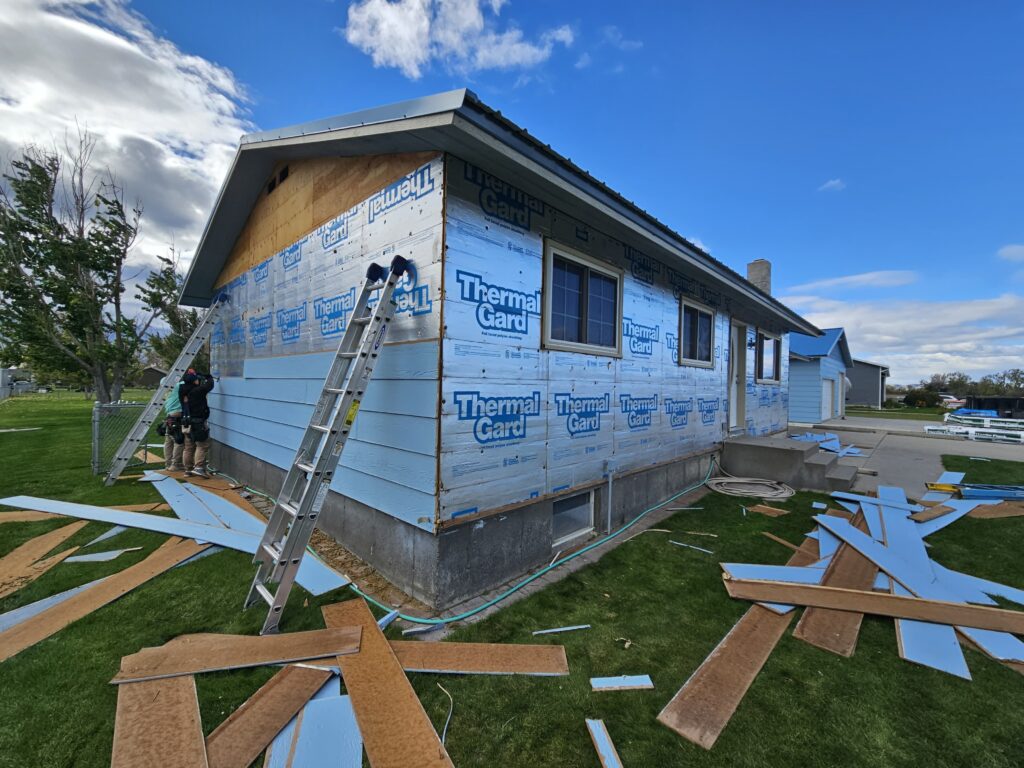Siding installation in progress by Nashville TN Siding Contractors Wegner Roofing & Solar. A house exterior is partially clad in blue Thermal Guard insulation board, with wooden siding partially installed on the front and workers on ladders completing the work under a blue sky.