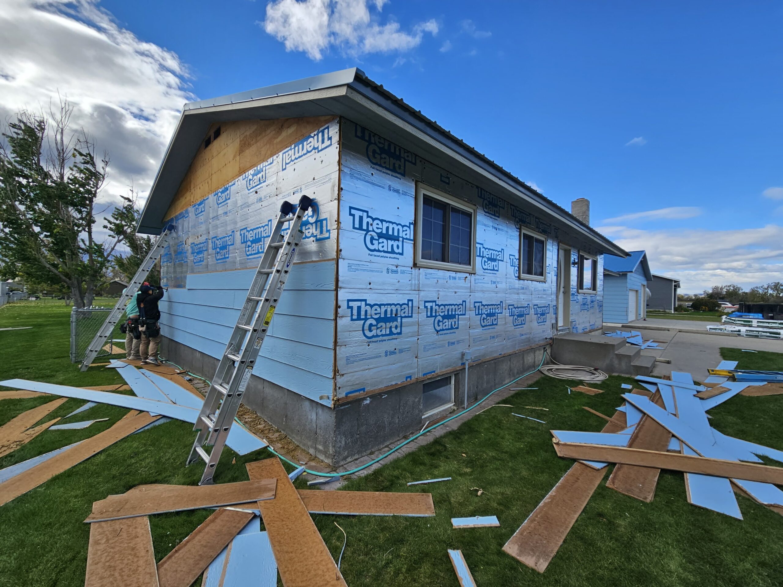Siding installation in progress by Nashville TN Siding Contractors Wegner Roofing & Solar. A house exterior is partially clad in blue Thermal Guard insulation board, with wooden siding partially installed on the front and workers on ladders completing the work under a blue sky.