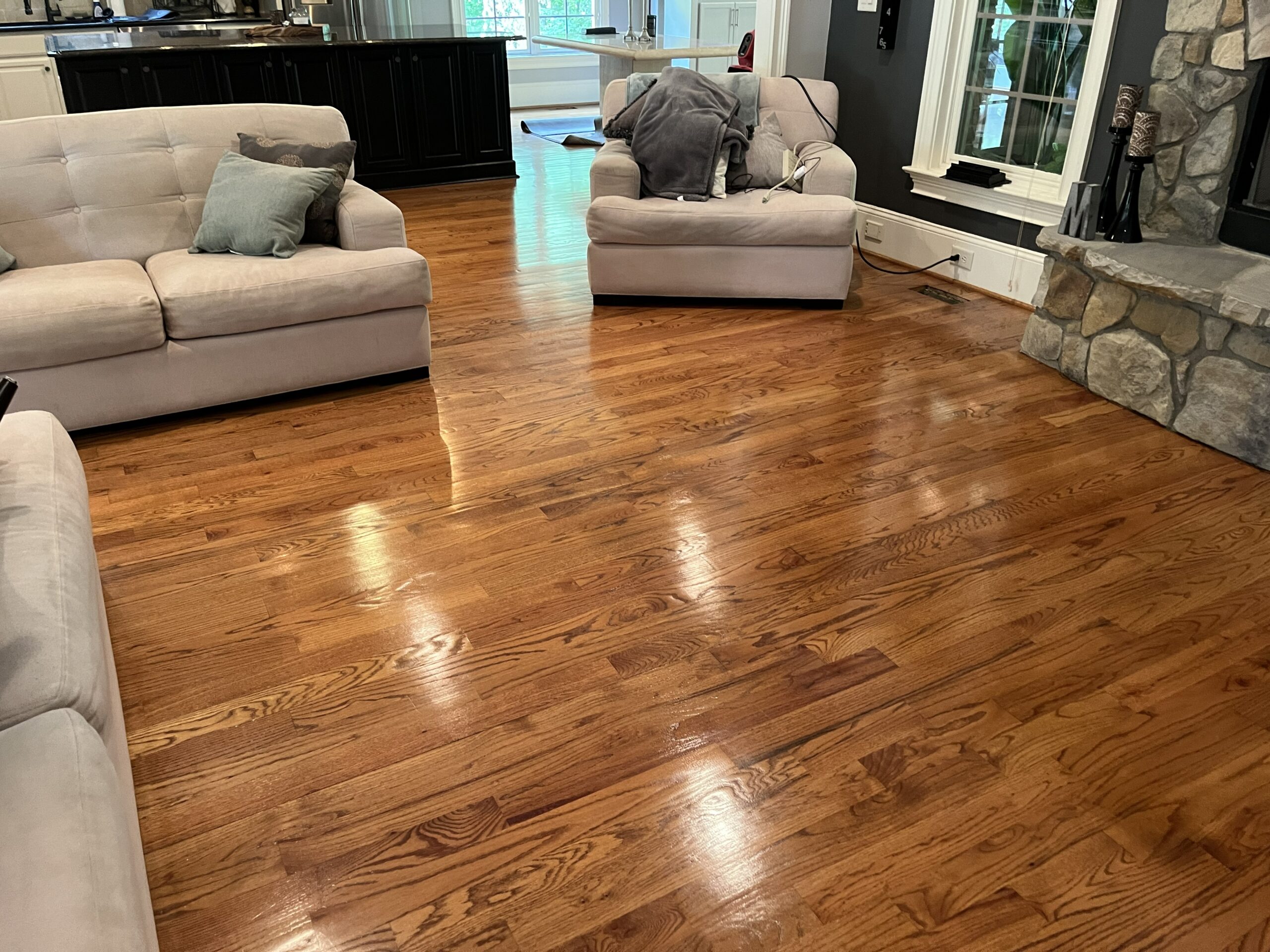 Newton NC Hardwood Floor Refinishing by Good House Floor Care. The image displays a newly refinished, glossy hardwood floor in a living area with light-colored sofas, a stone fireplace hearth, and a view into another room.