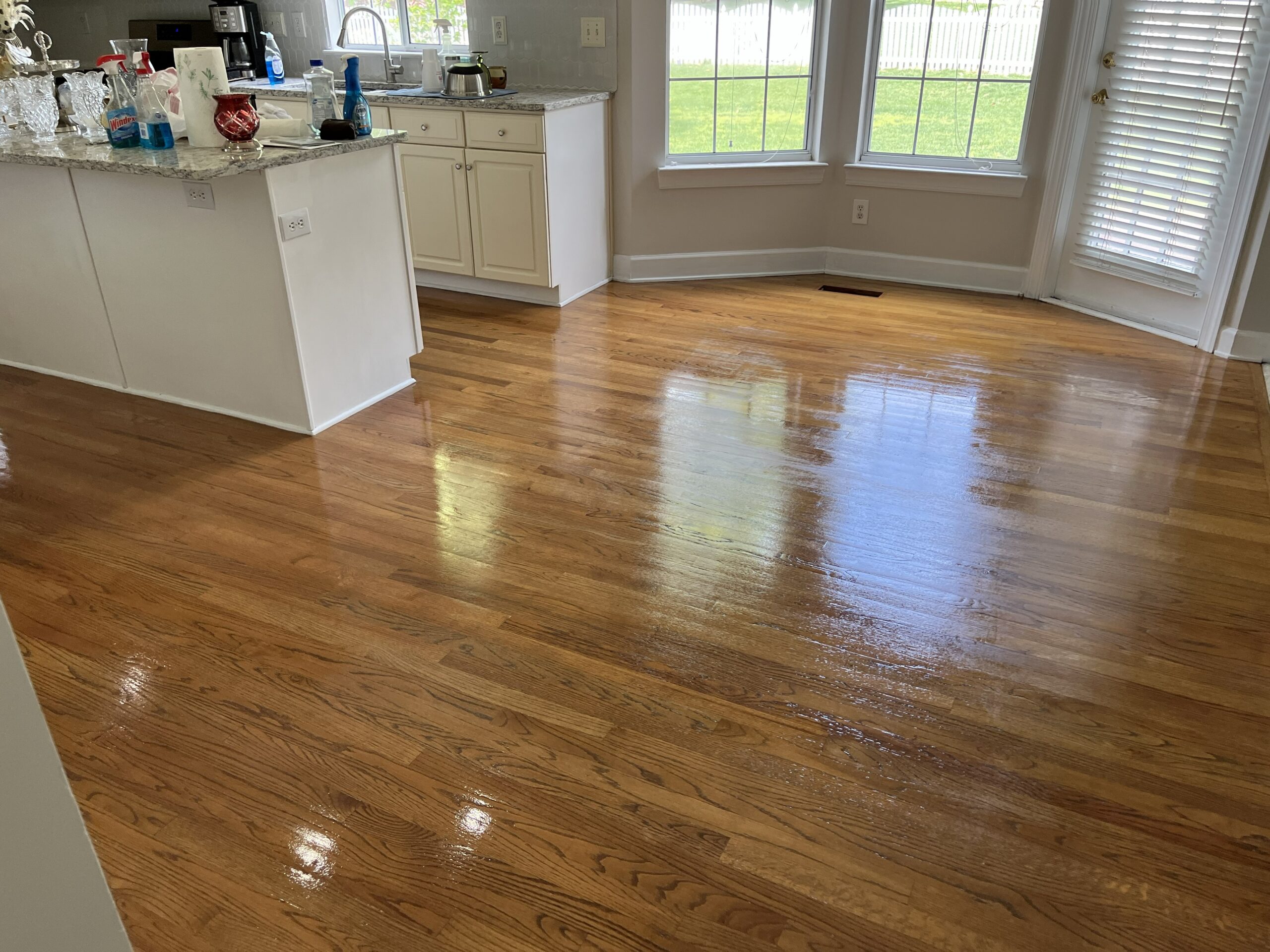 Newton NC Hardwood Floor Refinishing by Good House Floor Care. The image shows a newly refinished, glossy hardwood floor in a bright kitchen area with a white kitchen island, white cabinets, and windows in the background.
