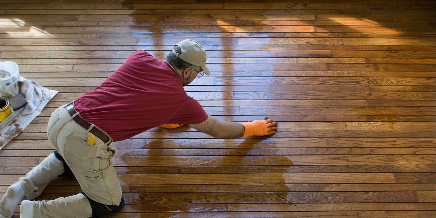 Contractor in a red shirt and cap on his knees applying a finish to a rich, dark brown hardwood floor, demonstrating the recoating step of "Newton NC Hardwood Screen and Recoat" by Good House Floor Care.
