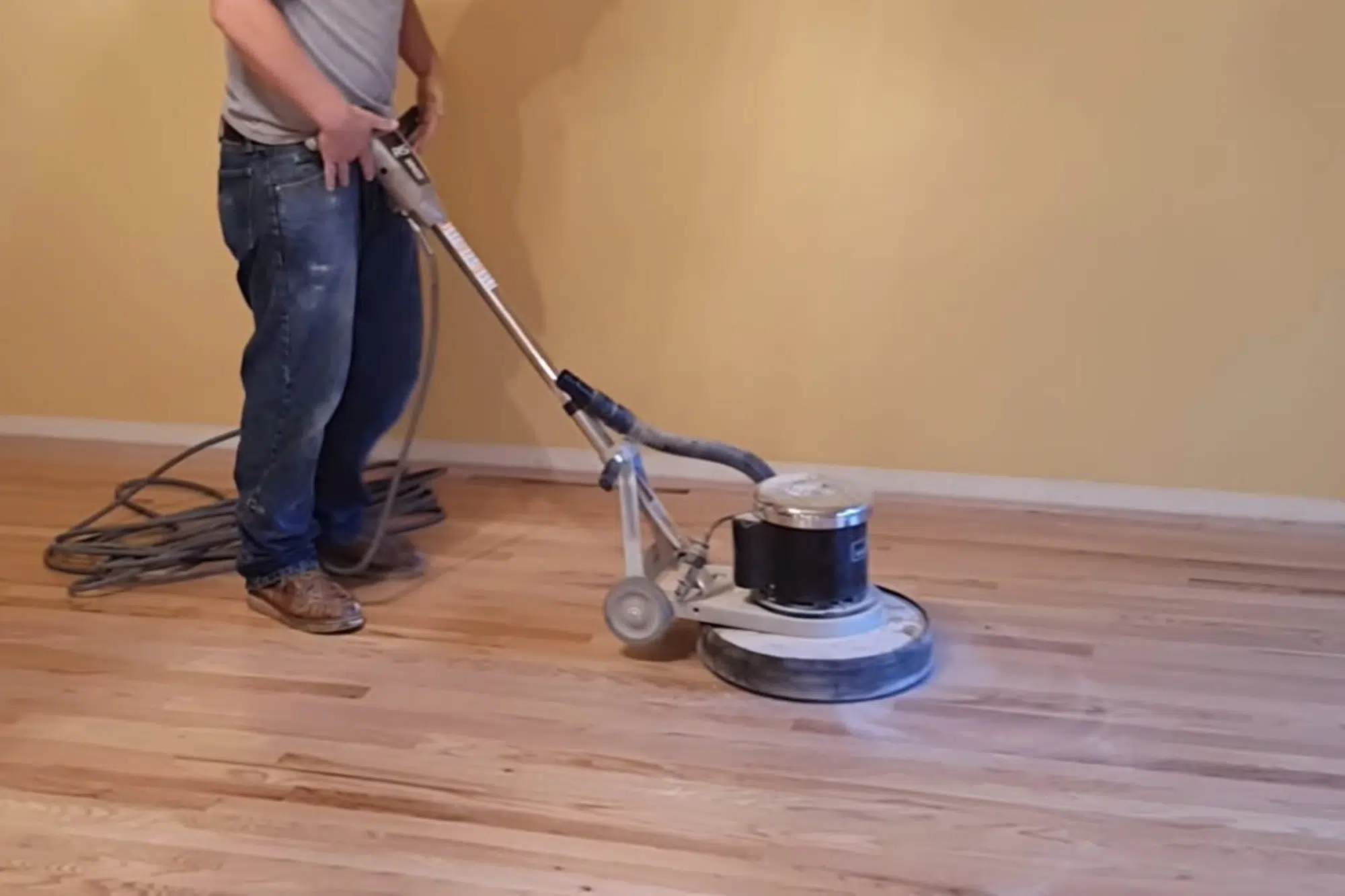 Man in jeans using a floor buffer machine on a light-colored hardwood floor as part of a "Newton NC Hardwood Screen and Recoat" service by Good House Floor Care.