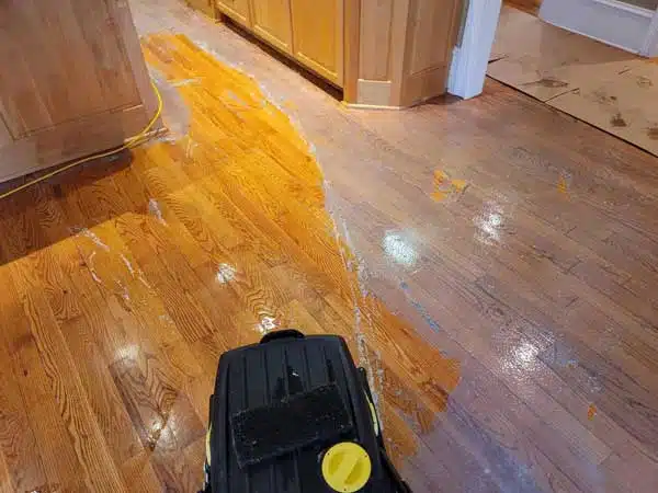 Wider view of a hardwood floor in a home in Newton, NC showing a clear delineation between the old, hazy wax coating and the freshly cleaned wood floor, with a wax removal machine visible. Service by Good House Floor Care.