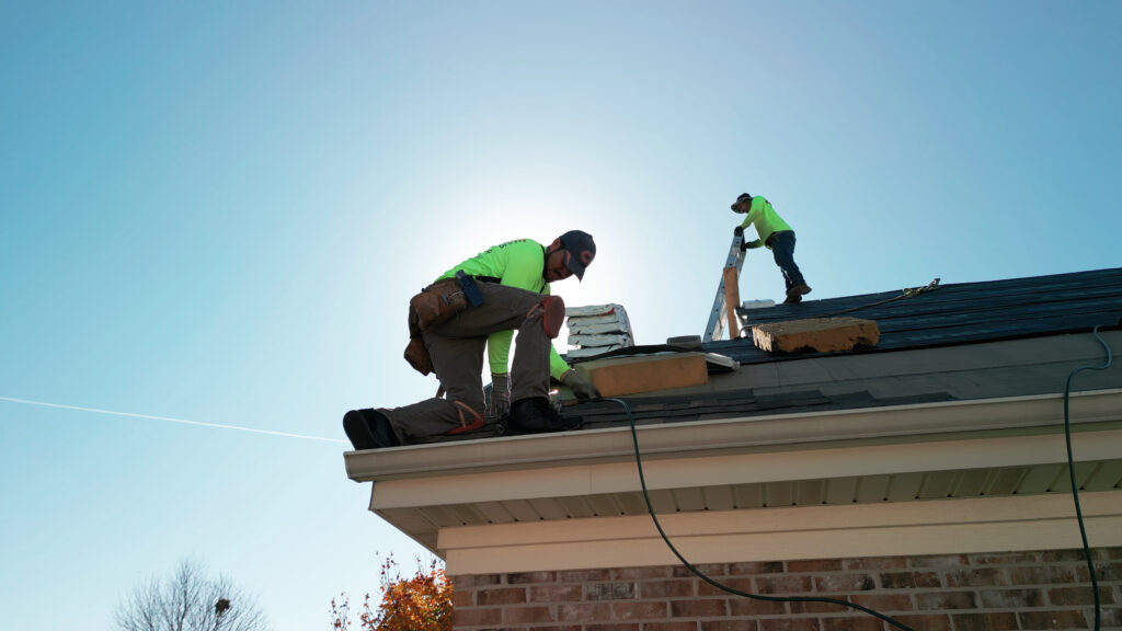 Two roofers working on a residential roof repair in North Palm Beach. One roofer is kneeling on the dark shingle roof, facing away from the camera, wearing bright green long-sleeve safety gear and a harness, with tools around his waist. The second roofer is further up the roof, climbing a ladder. The image is taken from a low angle against a bright blue sky, showcasing North Palm Beach Residential Roof Repairs by FL Pro Roofing & Solar.