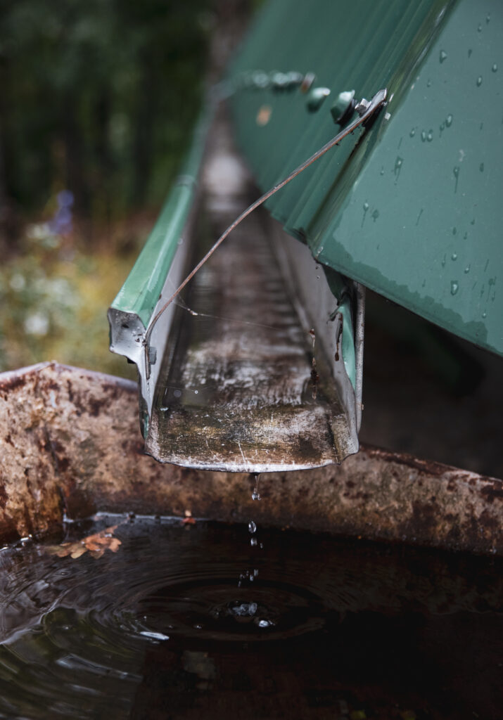Close-up of a damaged or overflowing green gutter system dripping water into a large rustic basin below, illustrating the need for Ramsey MN Gutter Damage repair by Pro24 Contracting, Inc.