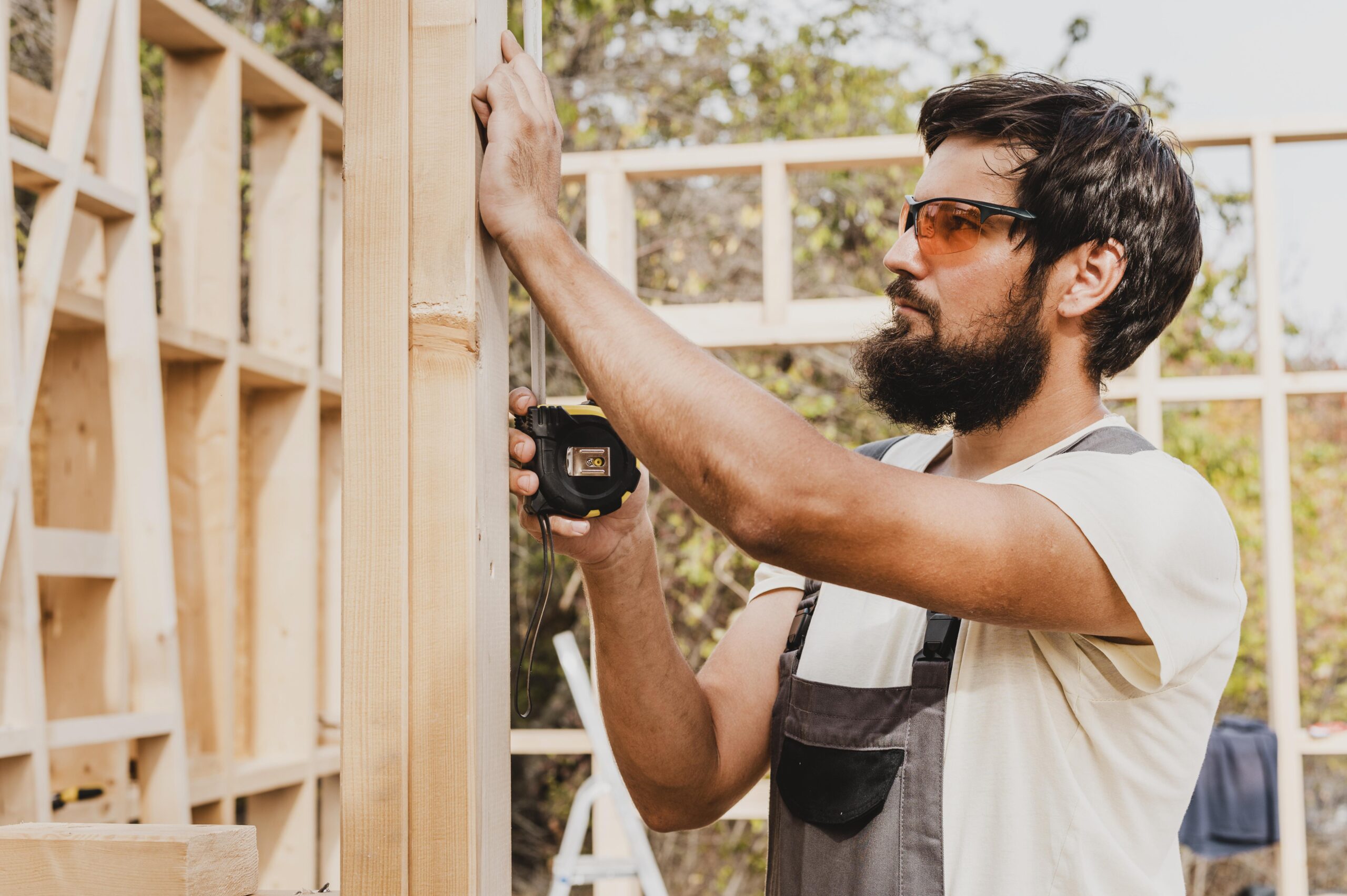 A bearded construction worker with safety glasses and overalls, identified as a "Ramsey MN Siding Contractor" by Pro24 Contracting, Inc., measures a wooden wall frame with a tape measure on a sunny day at a job site.