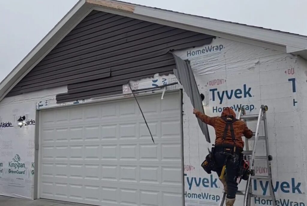 Close-up of a worker on a ladder installing Rapid City Vinyl Siding by Wegner Roofing & Solar on a home's gable. The worker, wearing an orange hooded top, is holding a section of brown vinyl siding against the house wrap, with partially installed siding visible above the white garage door.