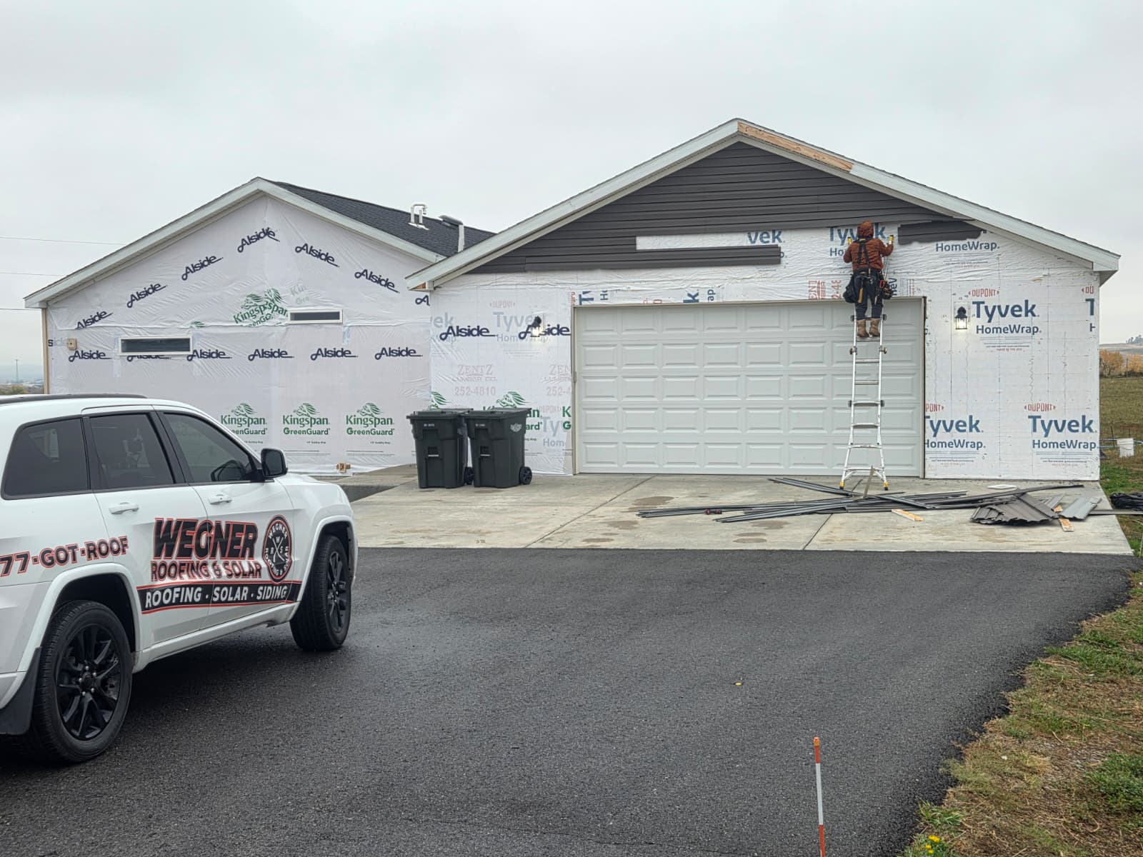 Installation of Rapid City Vinyl Siding by Wegner Roofing & Solar on a residential garage and home. The structure is wrapped in house wrap, and a worker is on a ladder installing brown vinyl siding on the front gable. A white Wegner Roofing & Solar branded vehicle is parked in the driveway.
