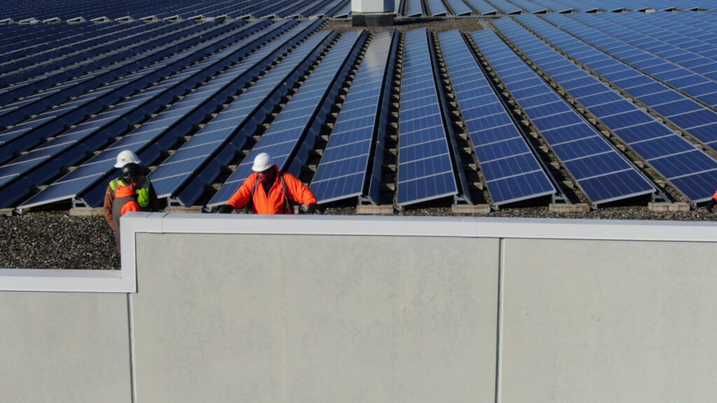 Close-up view of workers in safety gear (including a man in an orange jacket and white hard hat) standing near a low parapet wall on a commercial rooftop, with numerous rows of blue photovoltaic solar panels visible behind them, documenting the progress of the Wellington Solar Panel Installation by FL Pro Roofing & Solar.
