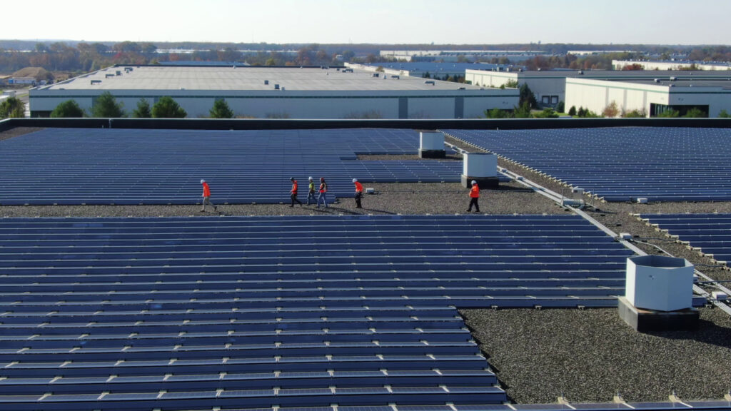 Wide aerial view of a massive commercial rooftop covered in rows of newly installed solar panels as part of the Wellington Solar Panel Installation by FL Pro Roofing & Solar. Several workers in orange safety vests and hard hats are walking across the solar array and the dark gray roof surface, with large warehouse-style buildings visible in the background under a partly cloudy sky.