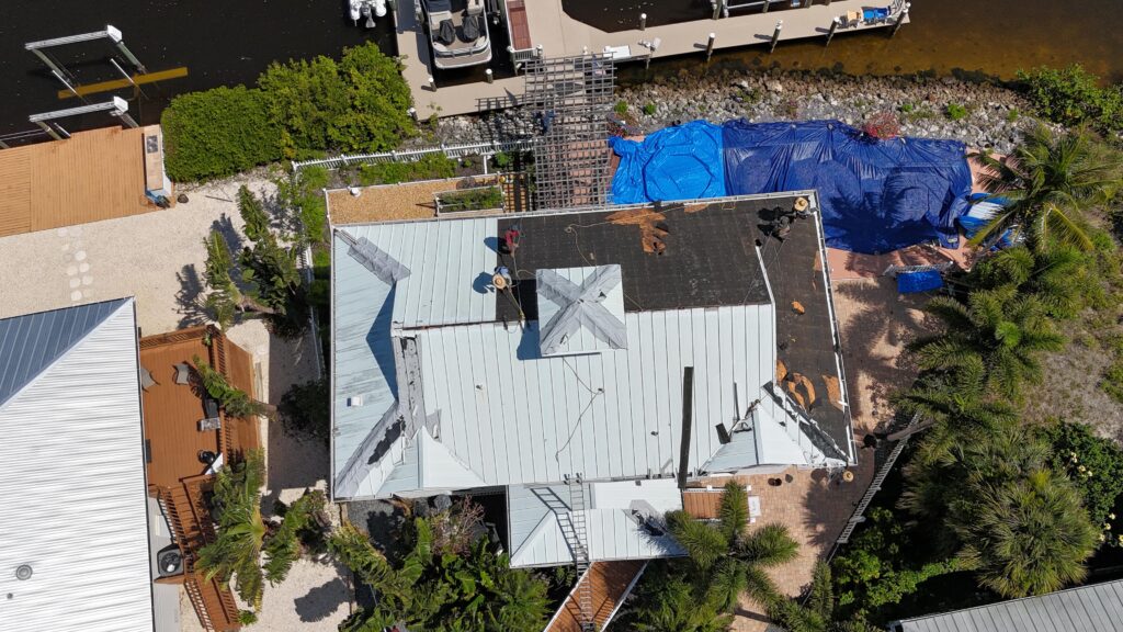 Drone view of an active metal roof tear-off and replacement project by a company among the top West Palm Beach Residential Roofing Companies by FL Pro Roofing & Solar. The image shows workers on a waterfront home, with the old black roofing partially removed and new light-colored metal panels being installed, and a blue tarp covering a section near the dock.