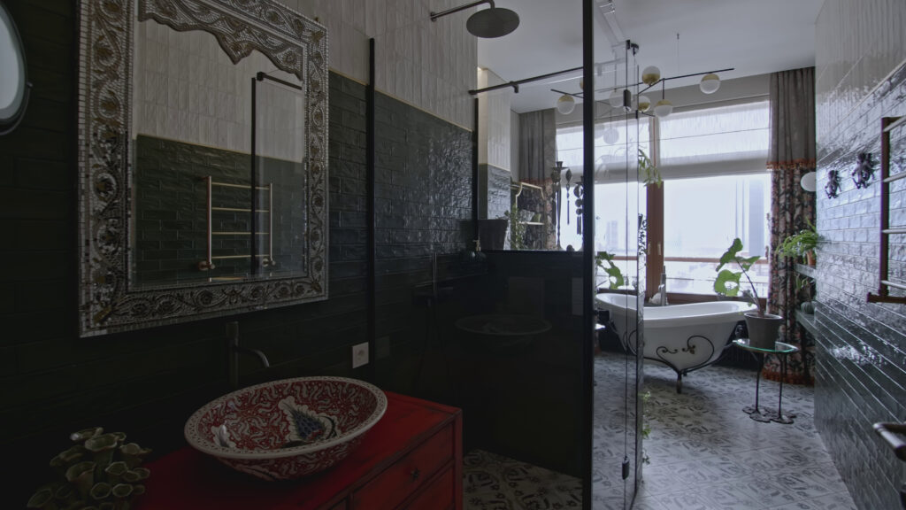 A luxurious, eclectic bathroom featuring a dark green subway-tiled shower area behind a glass partition, an ornate metal-framed mirror above a highly decorative red sink, and a white, claw-foot bathtub near a large window, showcasing high-end Aurora IL Shower Remodelling by Dream Bath Solutions.