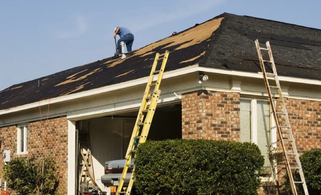 A roofer working on a residential roof replacement, demonstrating Bozeman Emergency Roofing Services by Wegner Roofing & Solar. The image shows the dark shingles partially stripped, with exposed roof decking, and two extension ladders propped against the gutter, suggesting ongoing repair work on a brick-faced home.