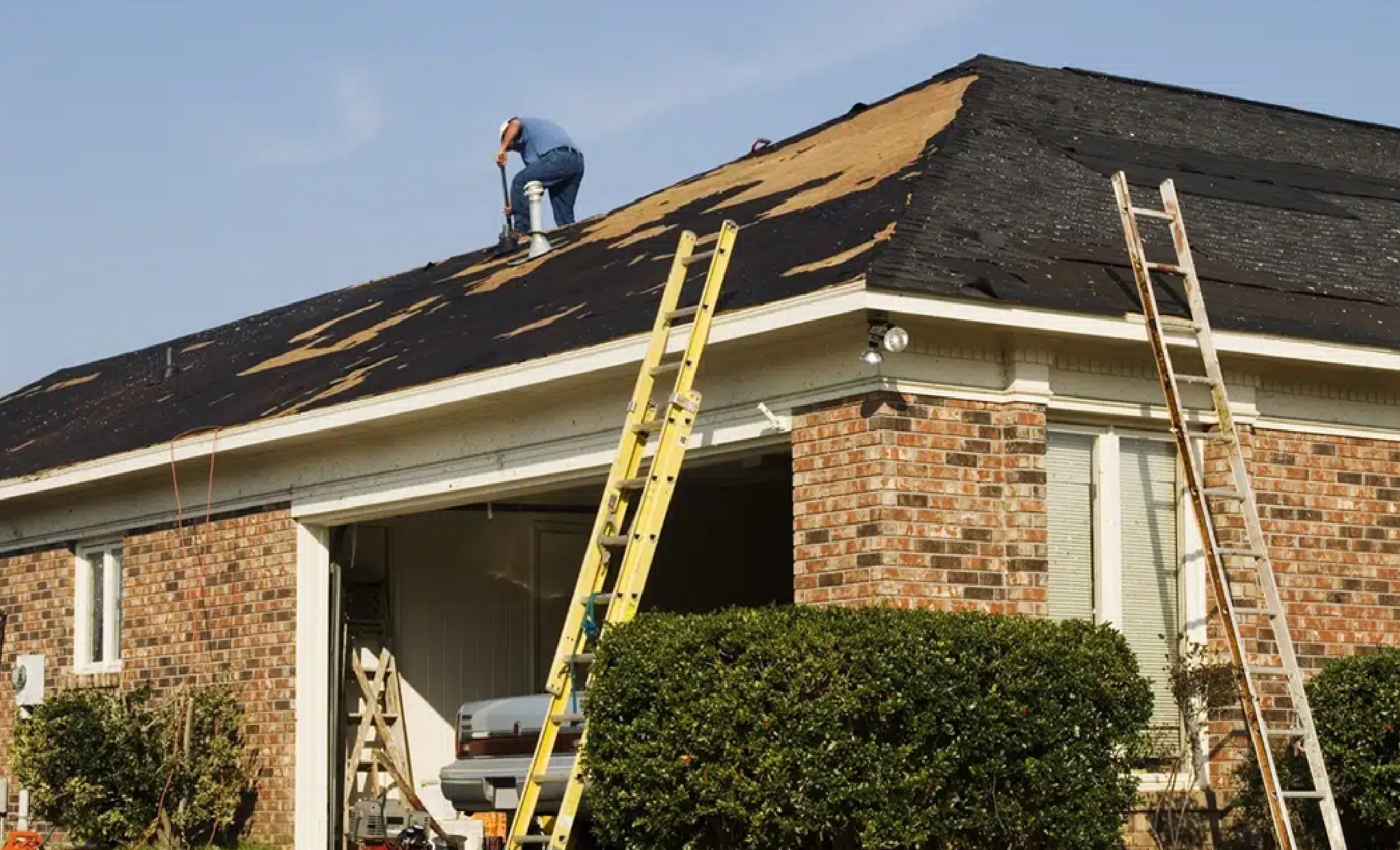 A roofer working on a residential roof replacement, demonstrating Bozeman Emergency Roofing Services by Wegner Roofing & Solar. The image shows the dark shingles partially stripped, with exposed roof decking, and two extension ladders propped against the gutter, suggesting ongoing repair work on a brick-faced home.